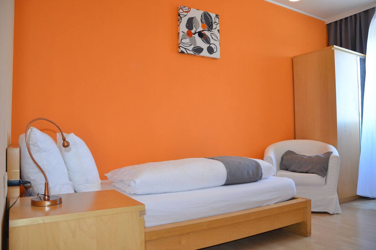 Un dormitorio con cama, mesita de noche, lámpara y una silla.