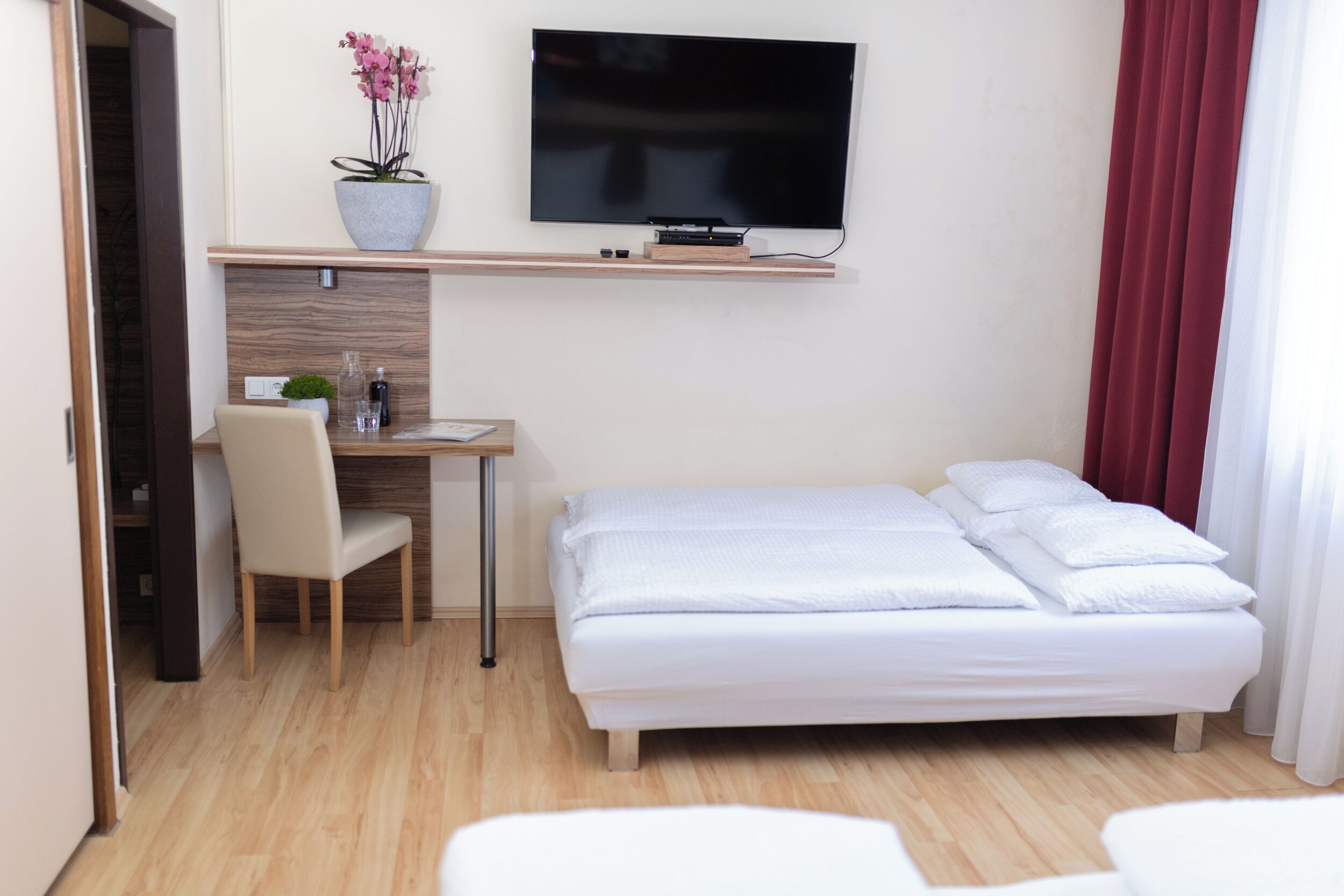 Une chambre d’hôtel comprenant un lit, un bureau avec une chaise, un téléviseur fixé au mur et une plante en pot.