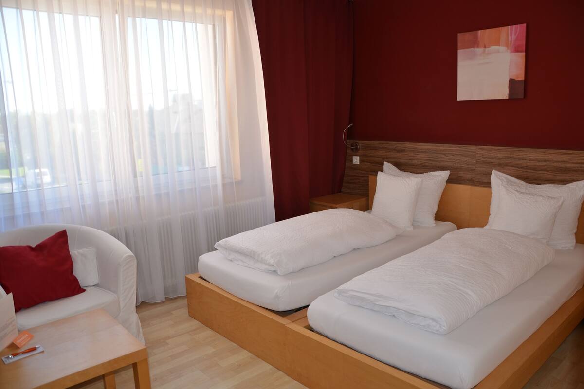 Habitación de hotel con dos camas individuales, cabecera de madera, sofá blanco con un cojín rojo, mesita de noche de madera y ventana con cortinas de gasa.