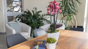 A wooden table with a plant and a book, surrounded by grey chairs in a room with a large window and potted plants.
