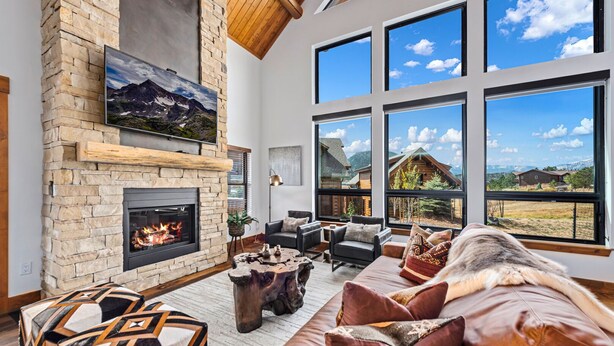 Amplia sala de estar con chimenea de piedra, grandes ventanales con vistas a un paisaje montañoso y muebles modernos.