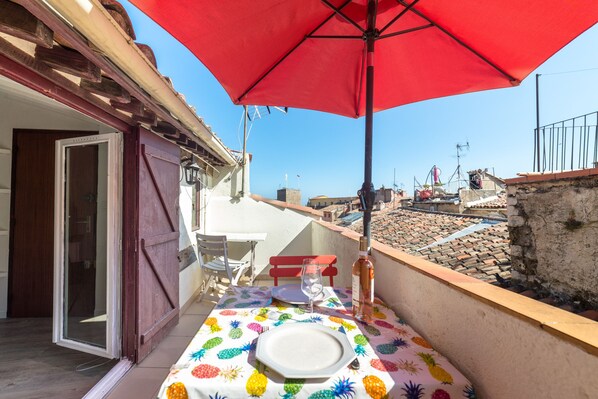 A balcony with a red umbrella, a table with a pineapple tablecloth, and a bottle of wine.