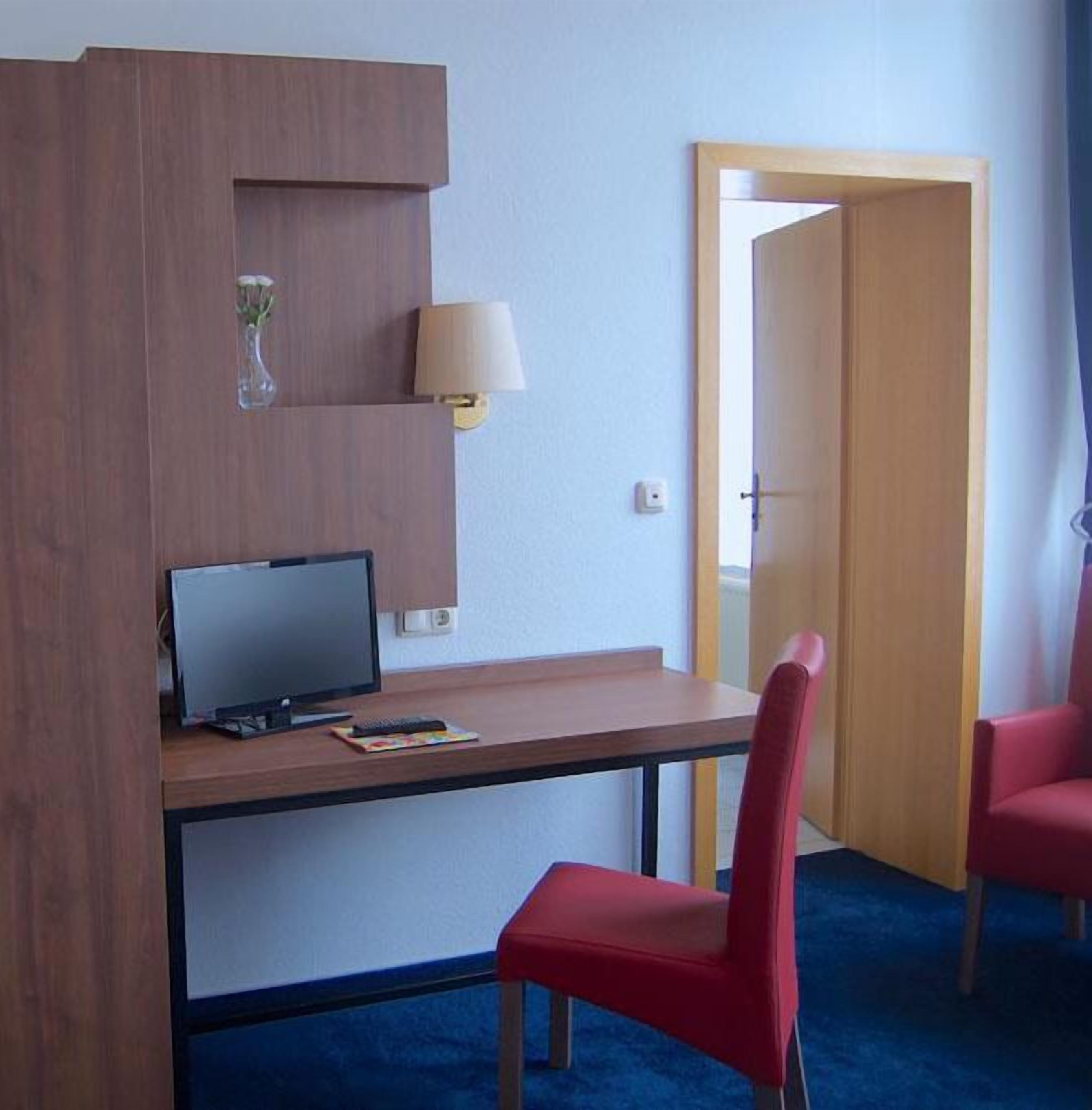 A hotel room with a wooden desk, a computer, a red chair, a lamp, and a mirror.