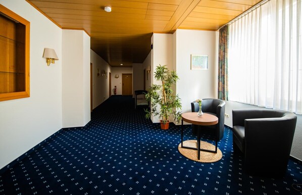 Un vestíbulo de hotel con una alfombra azul estampada, una mesa redonda con sillas, una planta en maceta y un gran ventanal con cortinas.