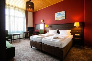 Una cama doble con ropa de cama blanca, un escritorio, una silla y un televisor en una habitación con paredes rojas y techo de madera.