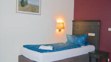 Una cama individual con colchón y almohadas azules, una mesita de noche, una lámpara de pared y un cuadro enmarcado.