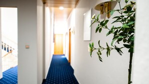 Een hotelgang met een blauw tapijt, witte muren en een plant aan de rechterkant.