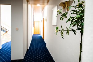 Un pasillo de hotel con alfombra azul, paredes blancas y una planta a la derecha.