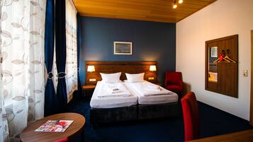 Habitación de hotel con dos camas, cabecera de madera, una silla roja, una mesa de madera y un cuadro enmarcado en la pared.