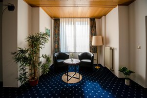 Una habitación de hotel con techo de madera, una gran planta de bambú en maceta, dos sillones de cuero negro, una pequeña mesa redonda con un jarrón, una lámpara de pie y un radiador.