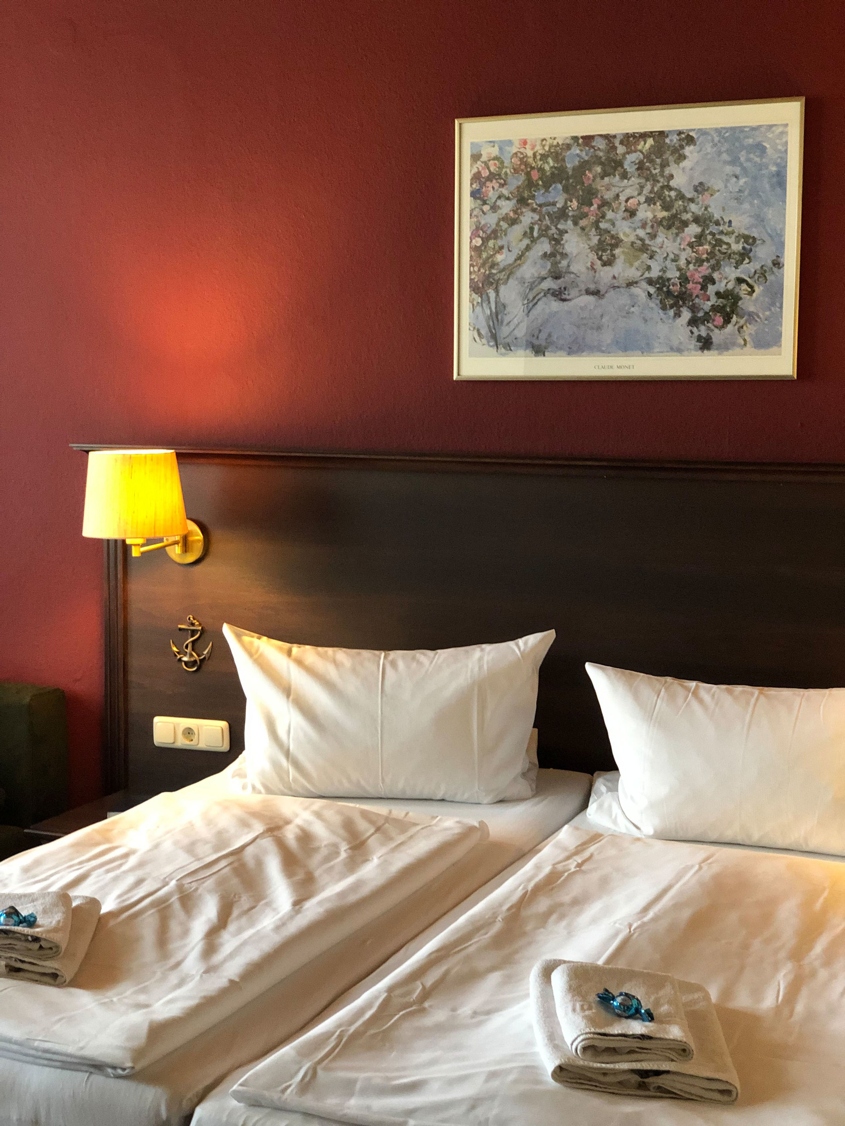 A hotel room with a double bed, white linens, a nightstand with a lamp, and a framed painting on the wall.