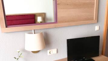 A hotel room with a wooden console table, a flat-screen TV, a lamp, a vase with a plant, and a mirror with a purple frame.