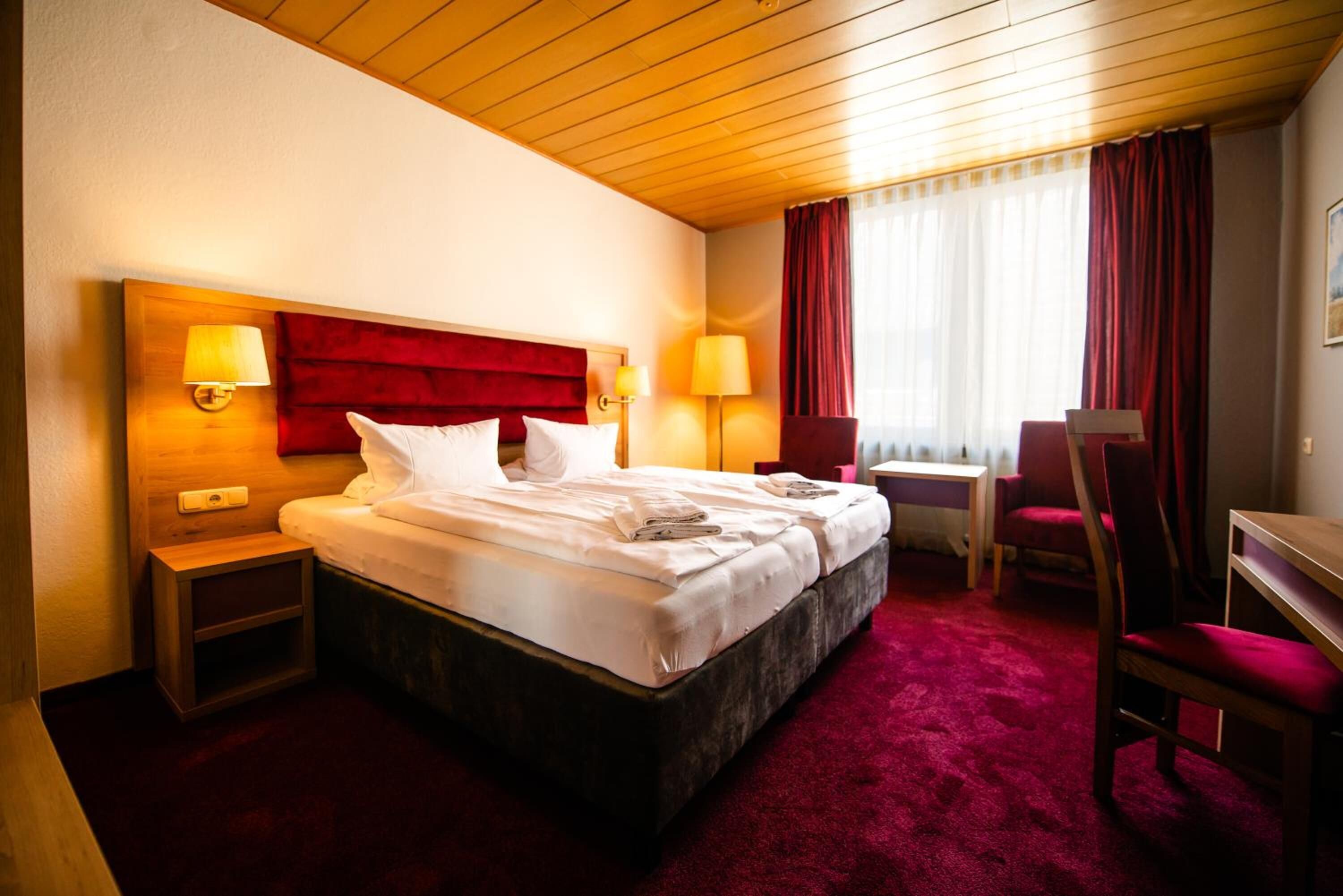 A hotel room with a large bed, two bedside tables, a desk, a chair, and a window with curtains.