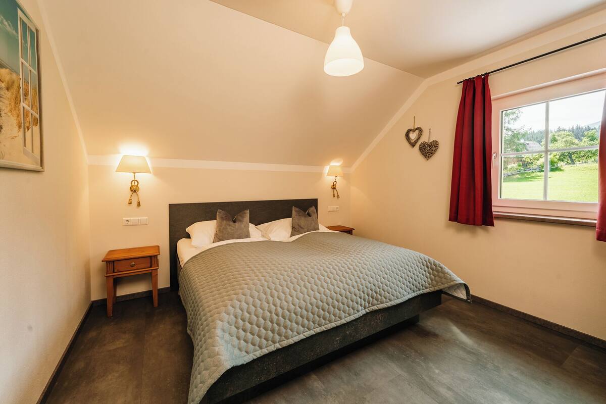 A bedroom with a bed, bedside lamps, a nightstand, a window with curtains, and a view of greenery outside.