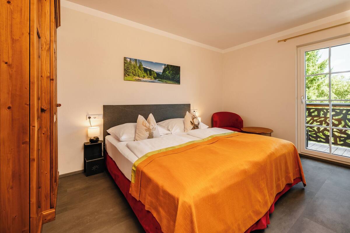 A bedroom with a bed, a nightstand, a red chair, a window with a view, and a wooden door.