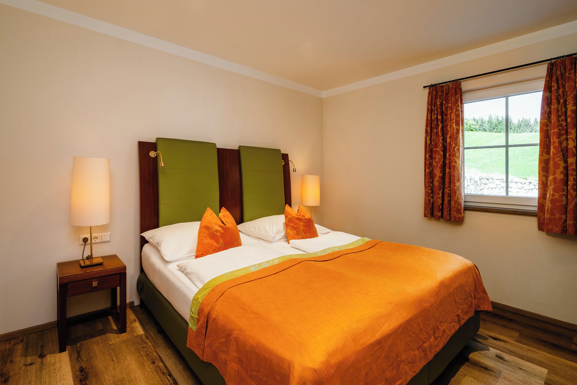 A hotel room with a bed, two bedside lamps, a nightstand, a window with curtains, and a view of greenery outside.