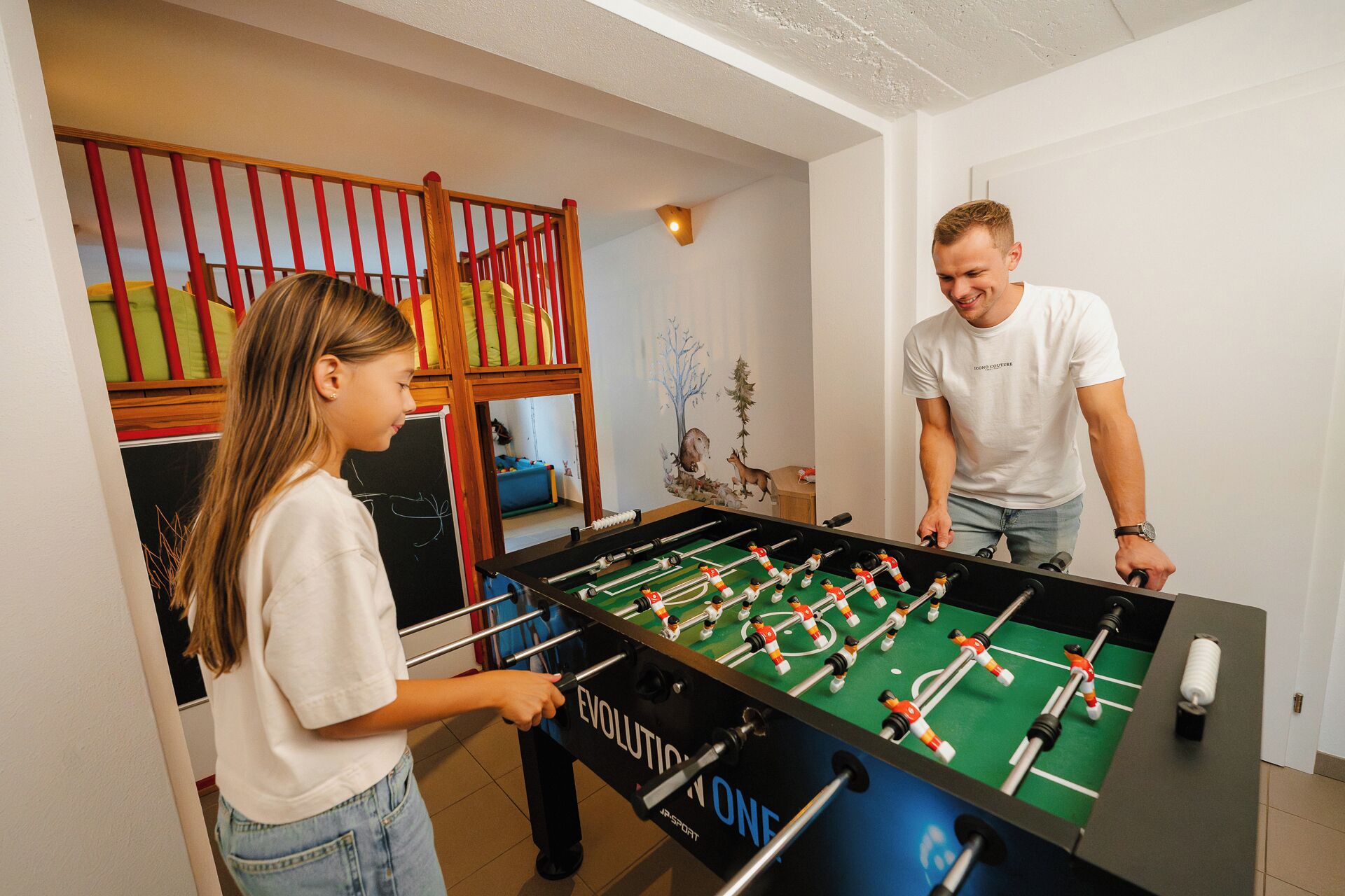 A room with a foosball table, a bunk bed with red and yellow bedding, and a wall with a mural.
