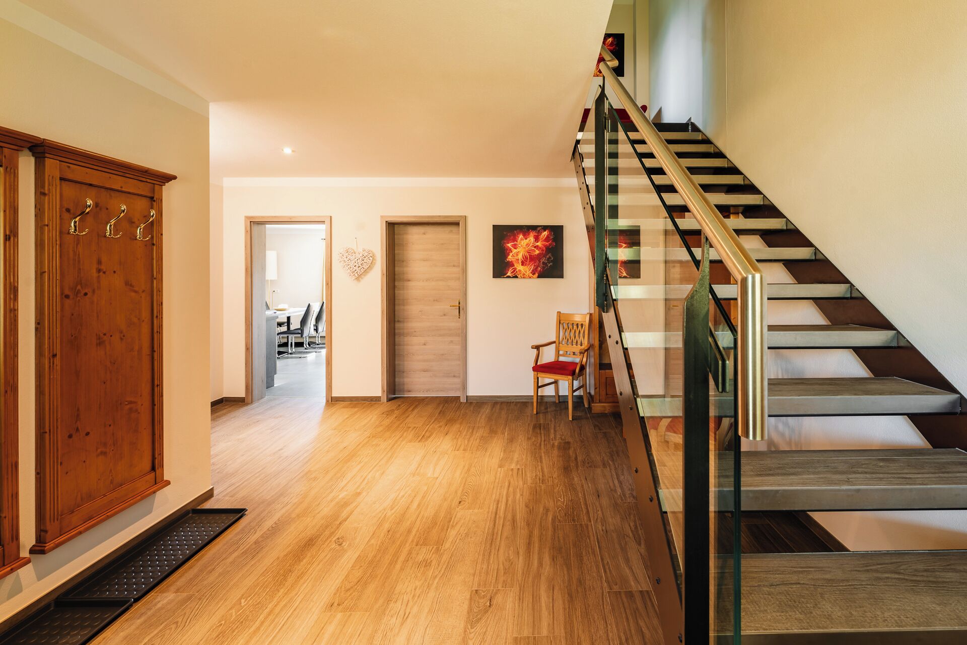 A staircase with a glass railing, wooden flooring, and a chair in the hallway.