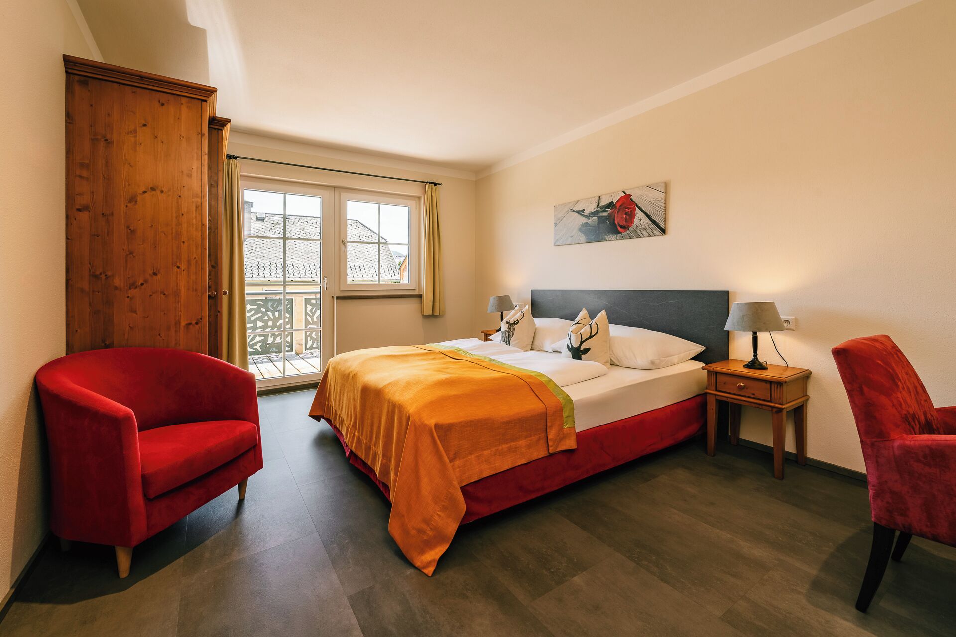 A bedroom with a bed, two red chairs, a wooden wardrobe, a nightstand with a lamp, and a window with a view.