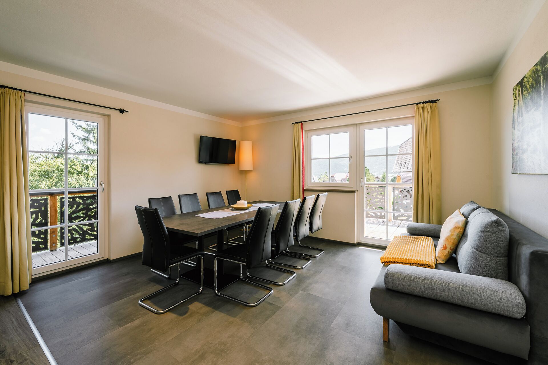 Ein Zimmer mit Esstisch, Stühlen, einer Couch, einem Fernseher und Fenstern mit Vorhängen.