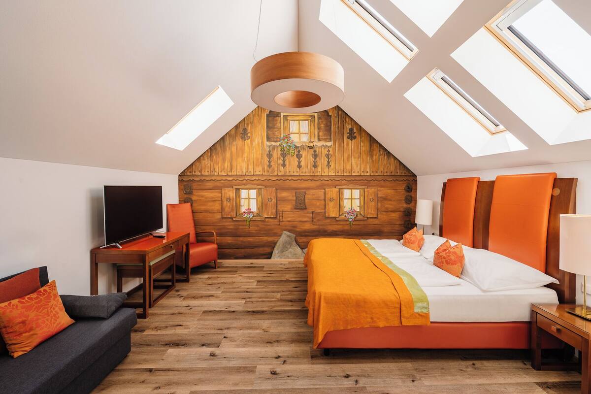 A bedroom with a sloped ceiling, wooden walls, a bed with orange bedding, a desk with a TV, and skylights.