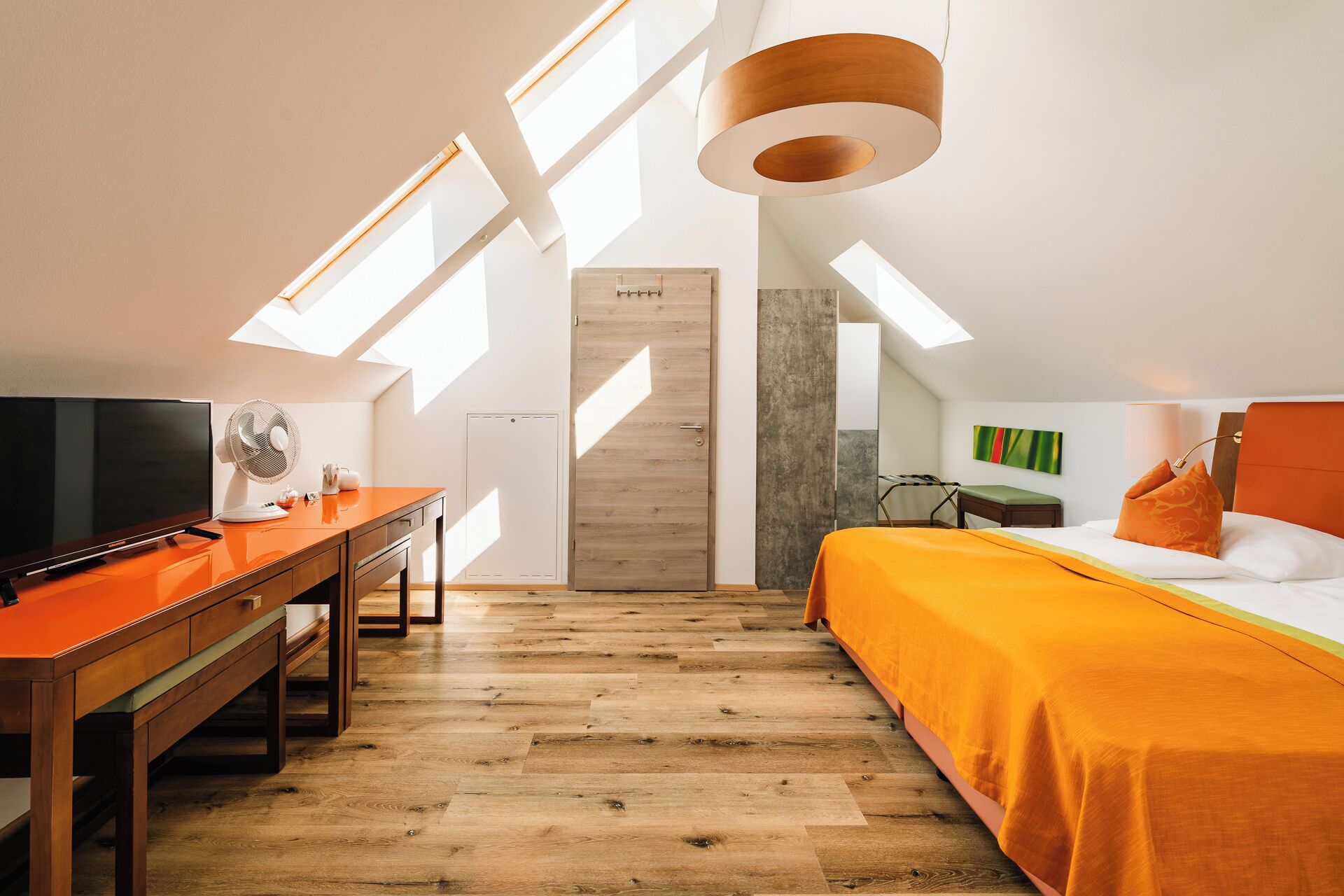 A modern hotel room with a wooden desk, a bed with orange bedding, a TV, and skylights.