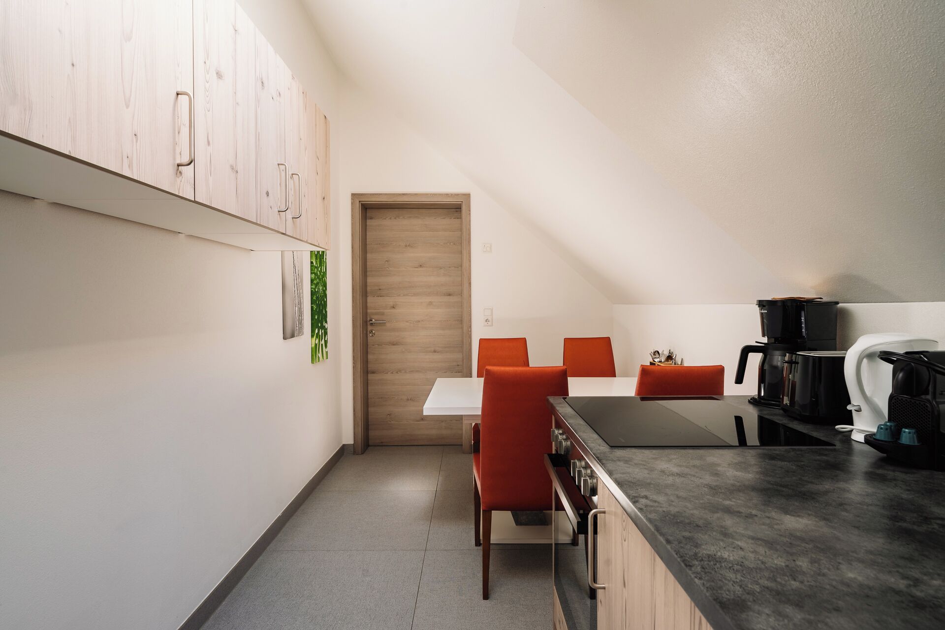 A compact kitchen with a dining area, featuring a wooden door, a white table, and orange chairs.
