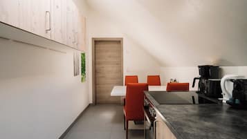 A compact kitchen with a dining area, featuring a wooden door, a white table, and orange chairs.