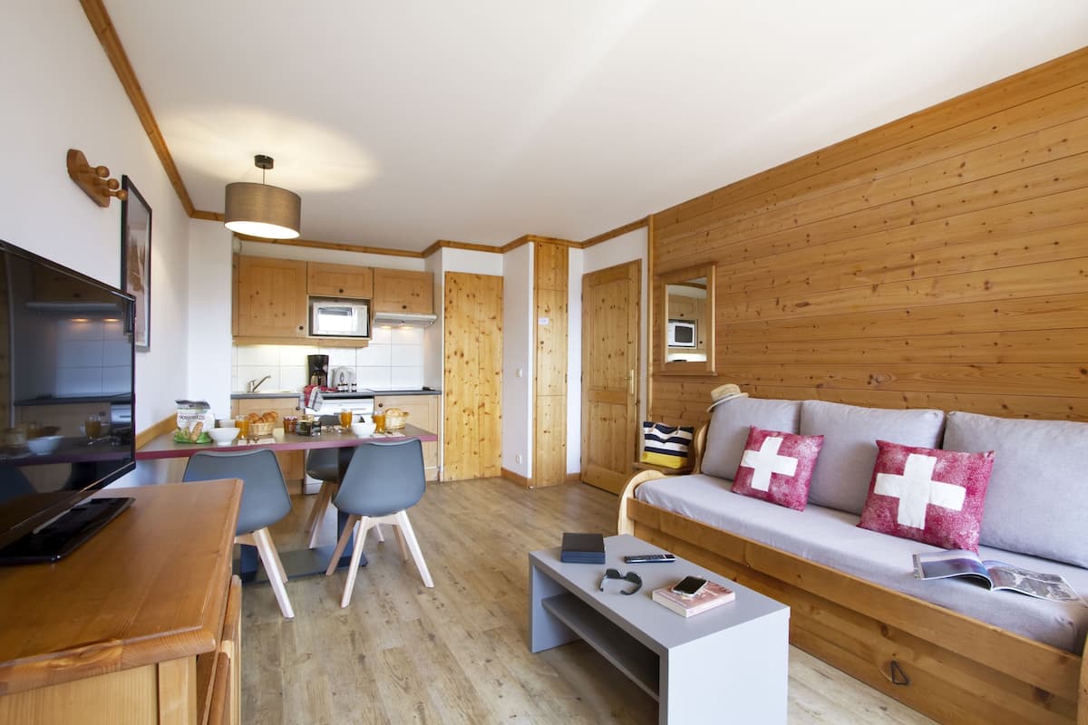 A modern living room with a wooden sofa, a dining area with a table and chairs, and a kitchen with wooden cabinets and a microwave.