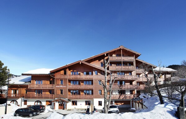 Ein mehrstöckiges Holzhaus mit Balkonen, im Hintergrund eine Skipiste und Schnee auf dem Boden.