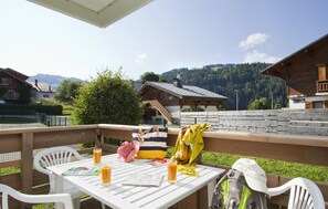 Eine Holzterrasse mit Tisch und Stühlen, ein gelber Pullover liegt über einem Stuhl, eine pinke Tasche und ein Fahrrad.