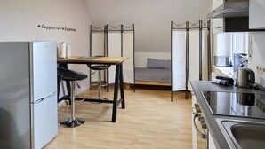 Een kleine keuken met koelkast, koffieapparaat, een tafeltje en een bed met matras en kussens.