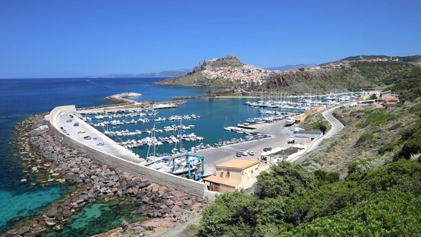 A coastal marina with boats docked, a stone wall, and a hill with buildings. - B&B Mimì (Castelsardo)