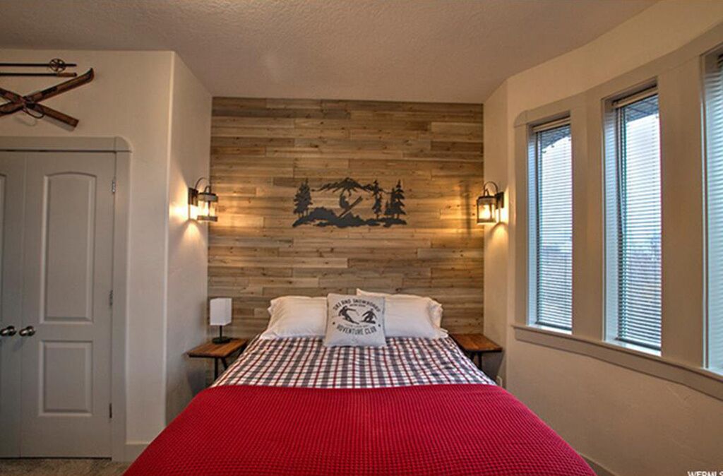 Ein Schlafzimmer mit einer Holzwand, einem Bett mit roter Decke, zwei Nachttischen mit Lampen und einem Fenster mit Jalousien.