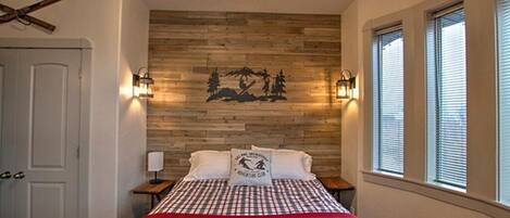 Ein Schlafzimmer mit einer Holzwand, einem Bett mit roter Decke, zwei Nachttischen mit Lampen und einem Fenster mit Jalousien.