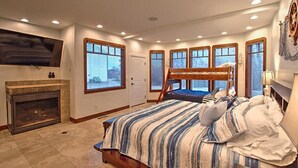 A bedroom with a fireplace, a large bed with a striped bedspread, multiple windows, and a ceiling with recessed lighting.