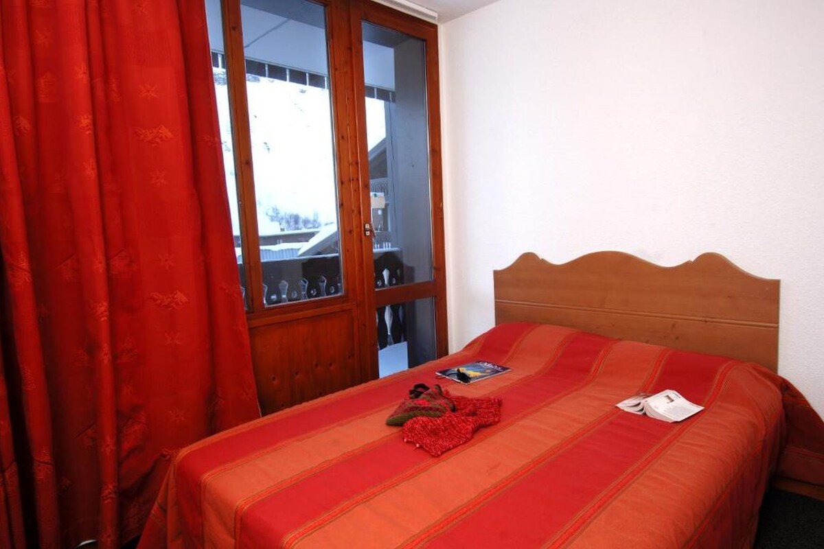 A bedroom with a wooden headboard, a bed with a red blanket, a red curtain, and a view through a glass door.