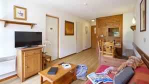 Un espacio habitable pequeño con un televisor en un mueble de madera, una mesita con un control remoto y un libro, un sofá con una manta y una zona de comedor de madera con sillas.