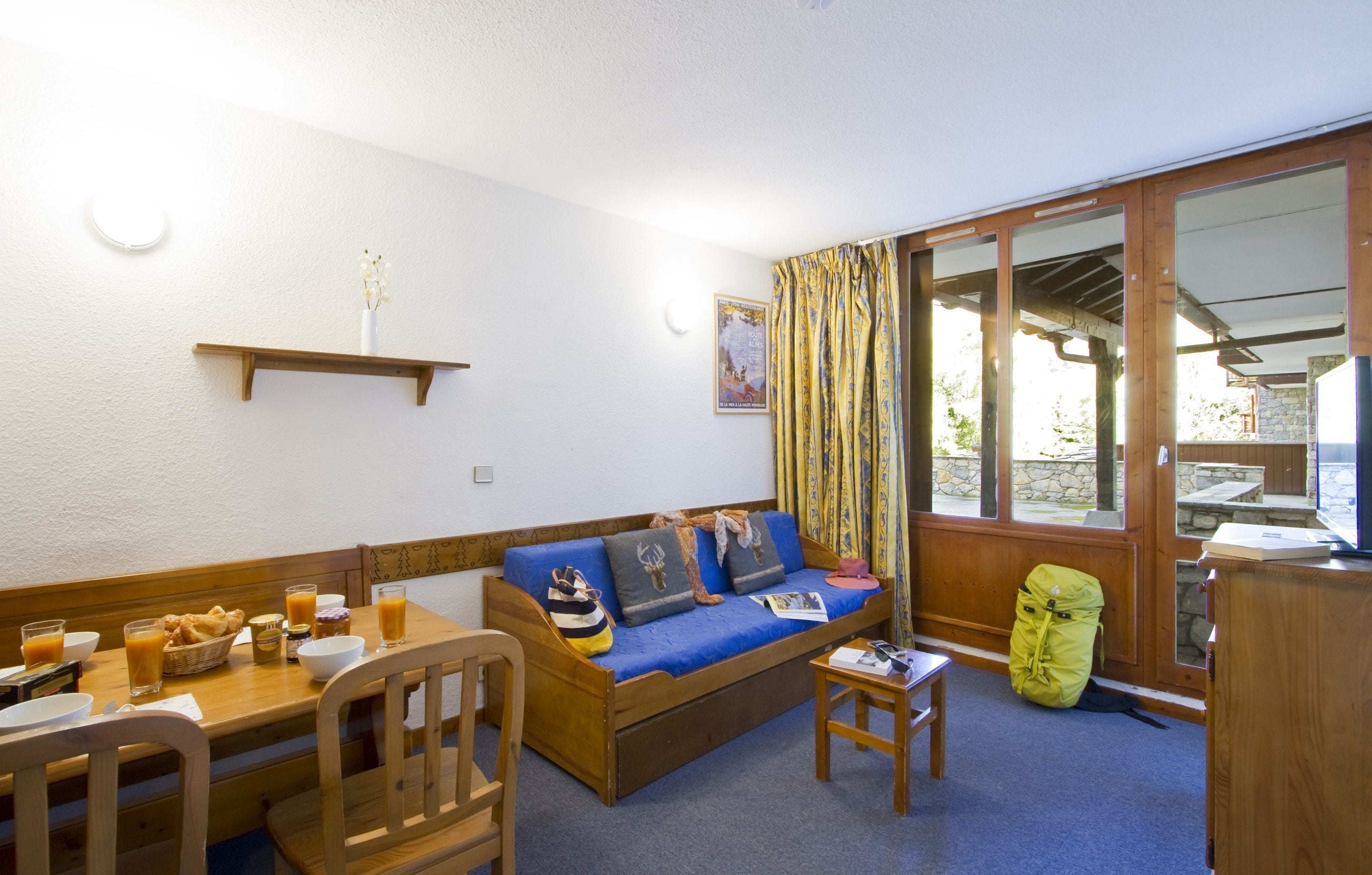 A cozy room with a wooden sofa, a dining table with chairs, a small table with a book, and a window with curtains.