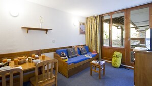 Una habitación acogedora con un sofá de madera, una mesa de comedor con sillas, una mesita con un libro y una ventana con cortinas.
