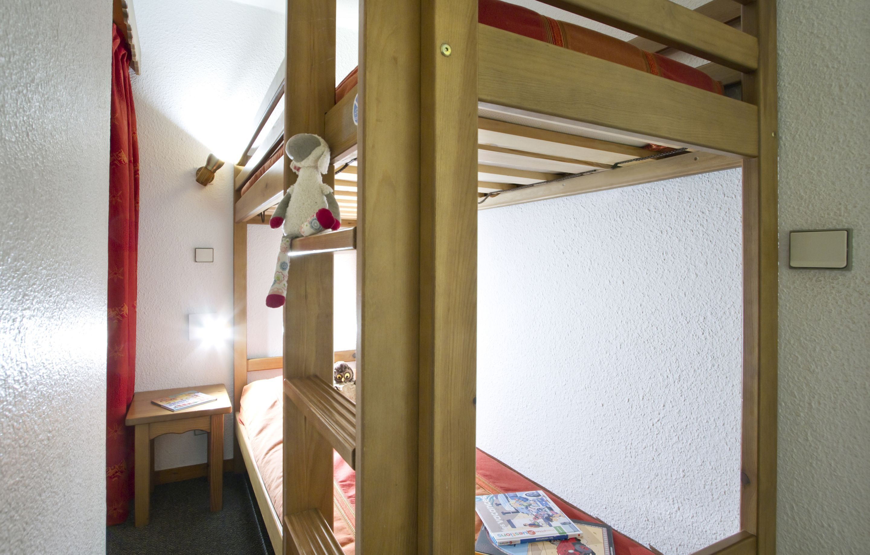 Una cama litera con una mesita pequeña y un libro, una cortina roja y un interruptor de luz en la pared.