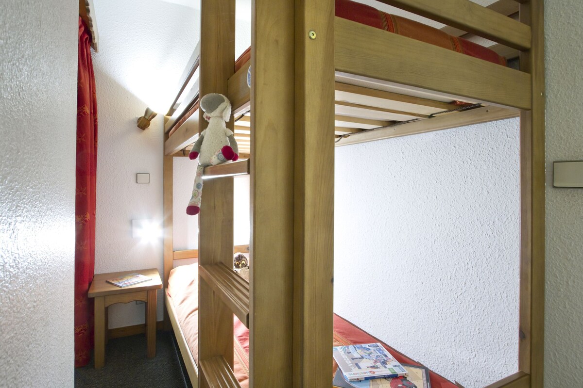 A bunk bed with a small table and a book, a red curtain, and a light switch on the wall.