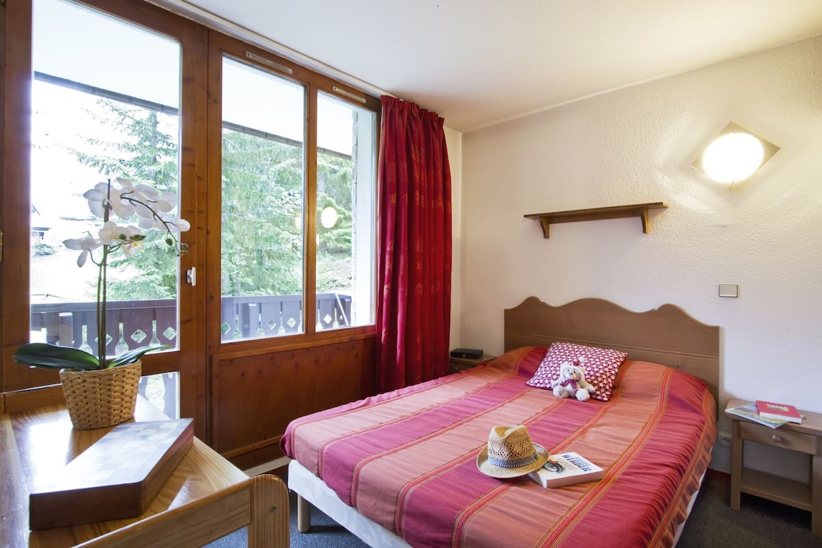 Um quarto com cama, mesa de cabeceira, cortinas na janela, cabeceira de madeira e prateleira na parede.