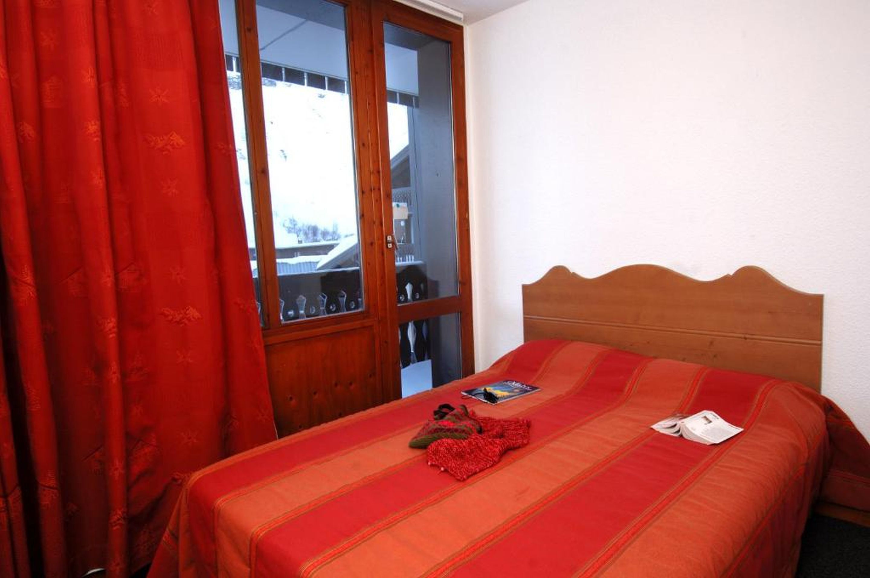 Un dormitorio con cabecera de madera, cama con manta roja, cortina roja y vista a través de un ventanal.