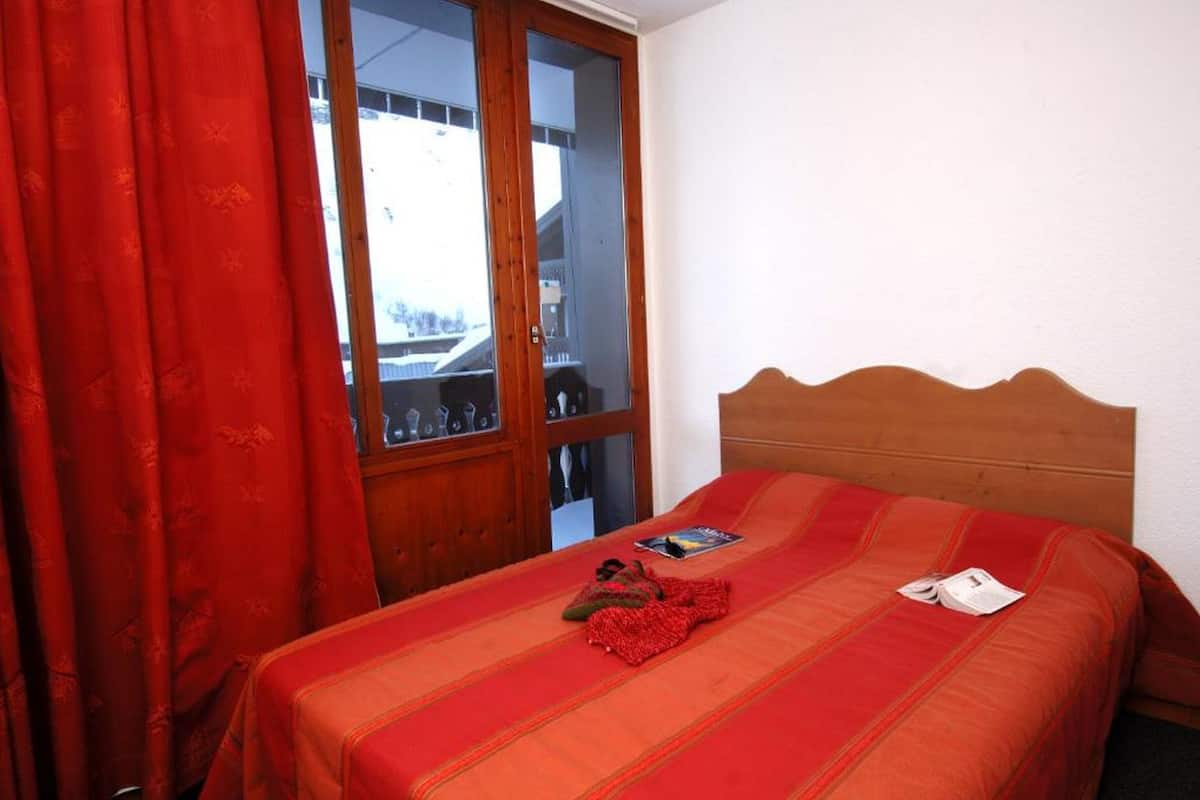 Quarto com cabeceira de madeira, cama com manta vermelha, cortina vermelha e vista por uma porta de vidro.