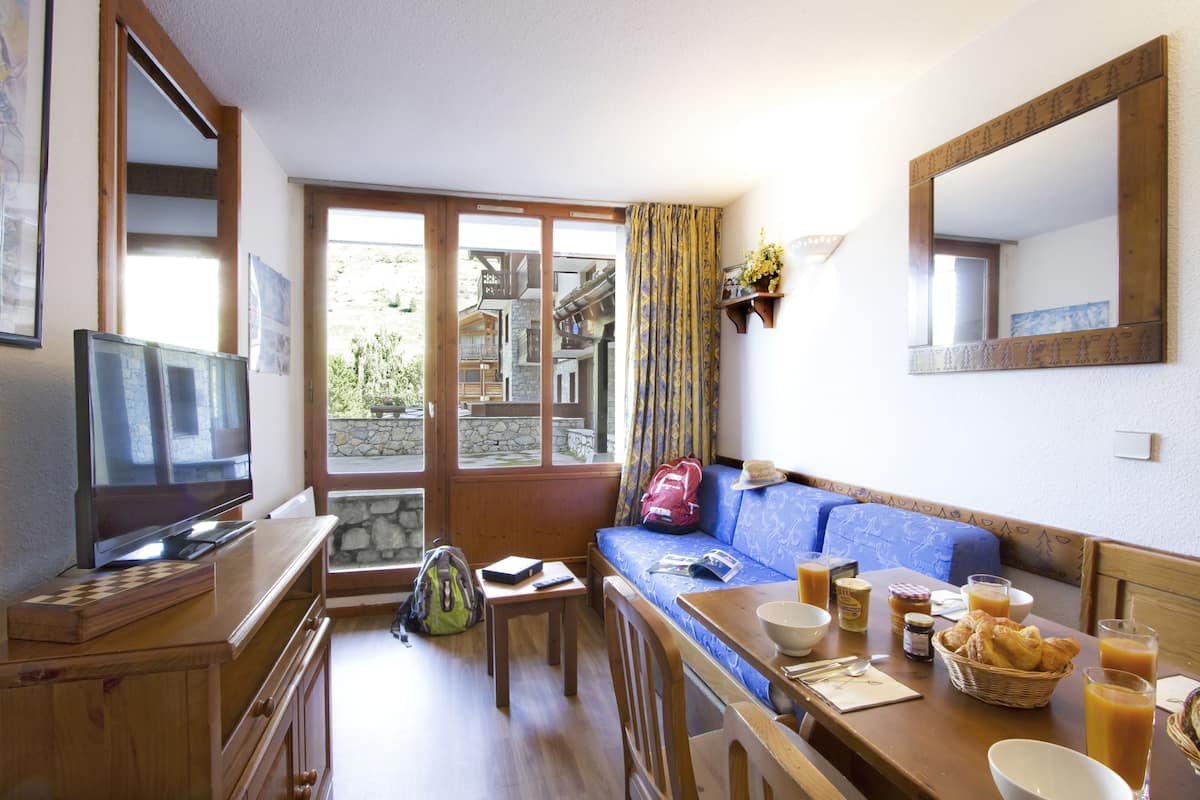 Um quarto aconchegante com um rack de madeira para TV, um sofá, uma mesa de jantar com itens de café da manhã e uma grande janela com vista para um edifício de pedra.