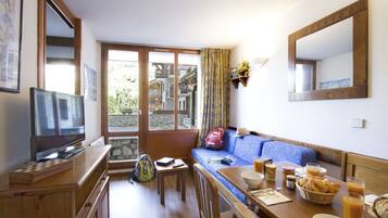 A cozy room with a wooden TV stand, a sofa, a dining table with breakfast items, and a large window offering a view of a stone building.