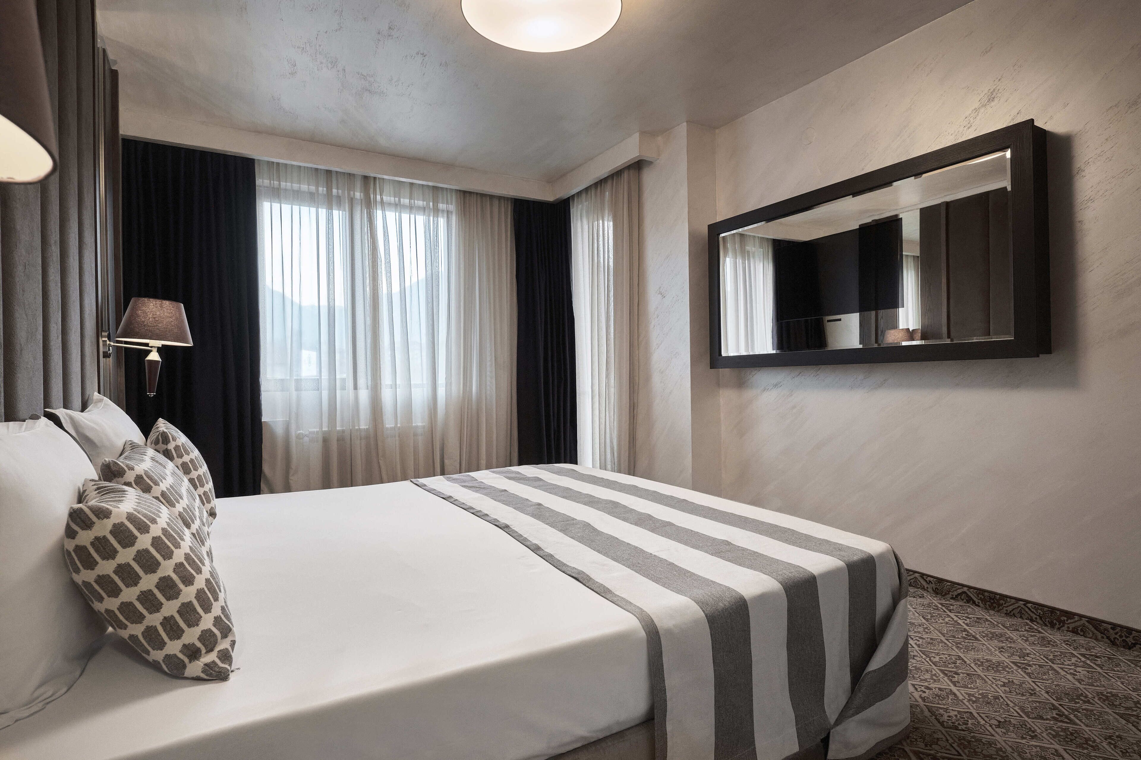 Habitación de hotel con una cama grande, una lámpara de noche, un espejo y vistas al exterior por la ventana.