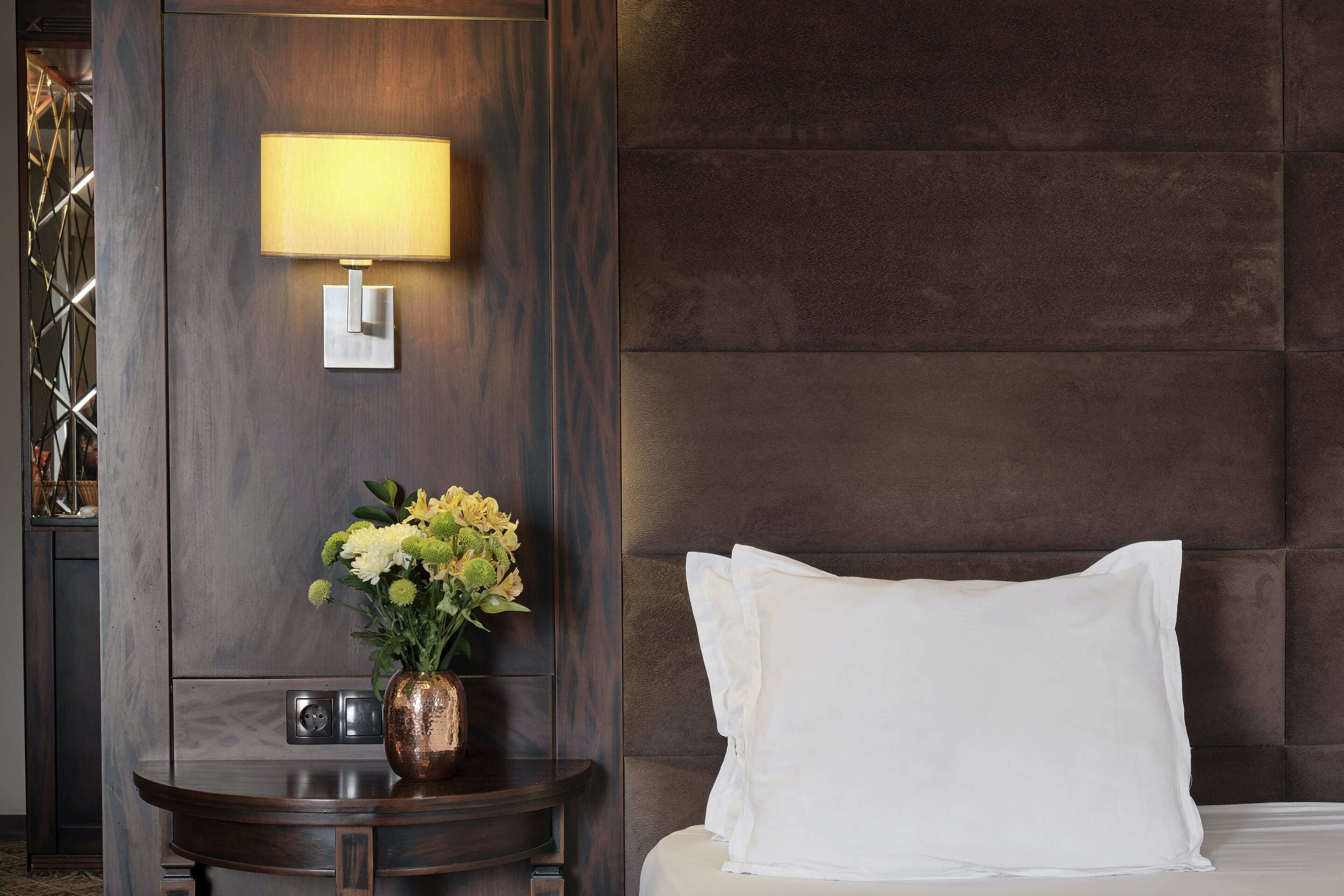 A hotel room with a bed, a bedside lamp, a vase with flowers, and a nightstand.