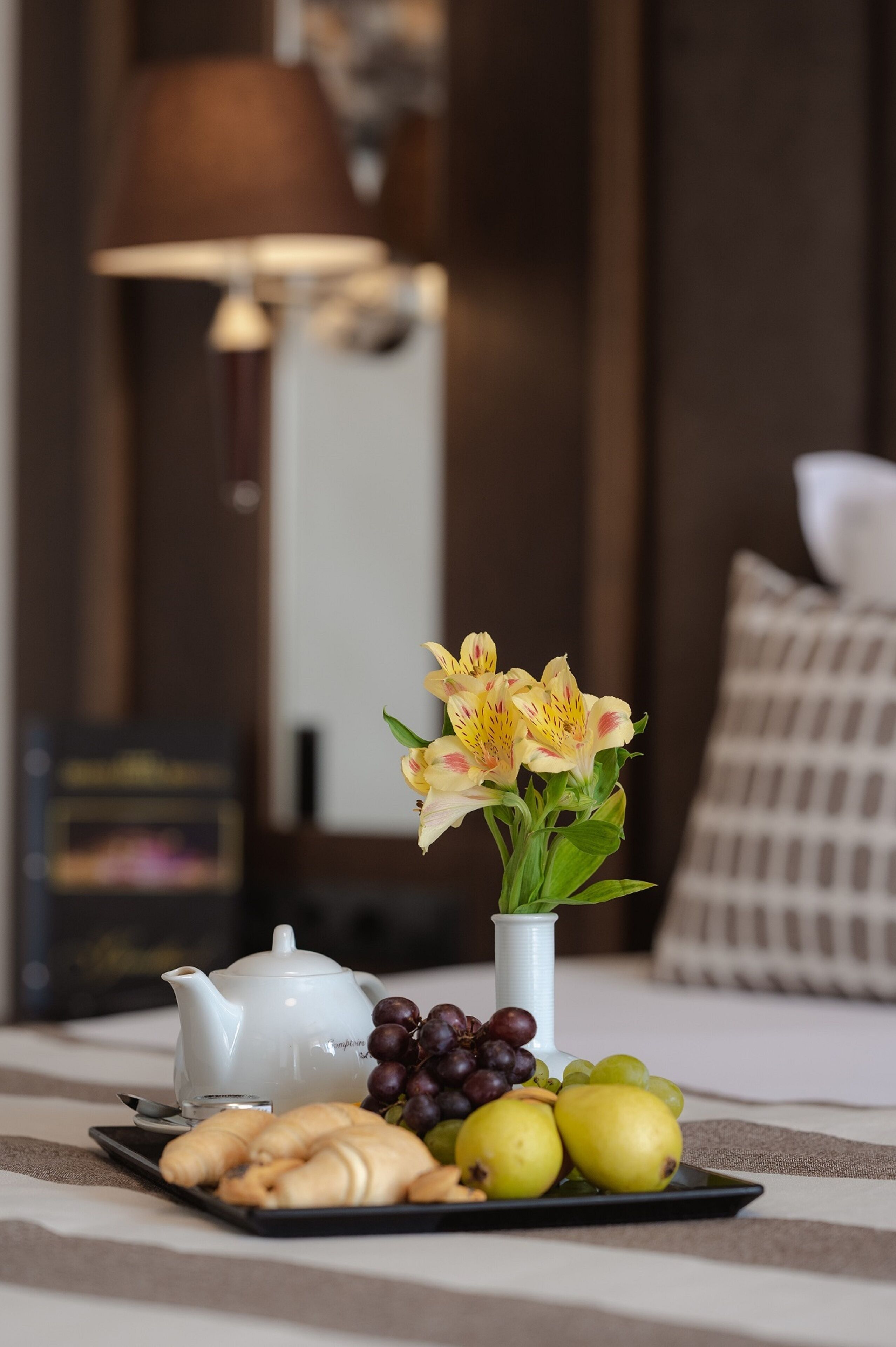 A tray with croissants, grapes, and pears on a table, a white teapot, and a vase with flowers in a hotel room.